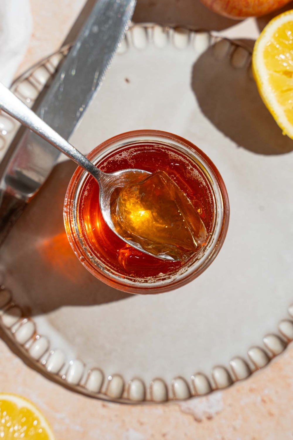 A glass jar of apple jelly with a spoon in the jelly. The jar is on a plate on a tan counter with a knife and sliced lemon.
