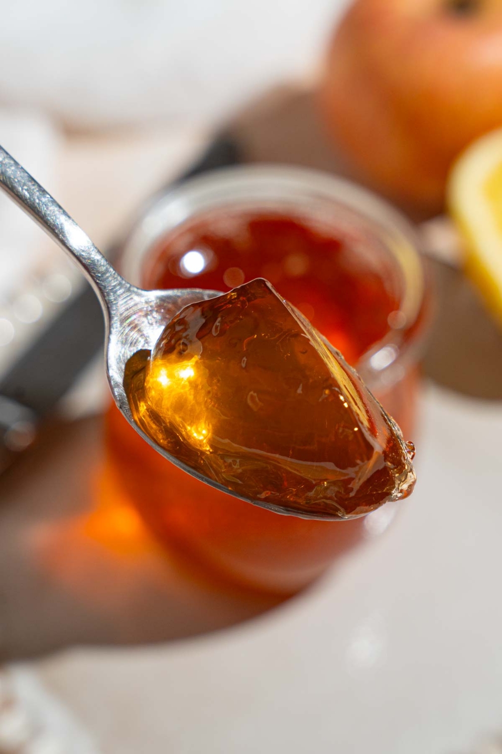 A close up of a spoon with apple jelly. There is a glass jar of jelly on a plate on a tan counter blurred in the background.