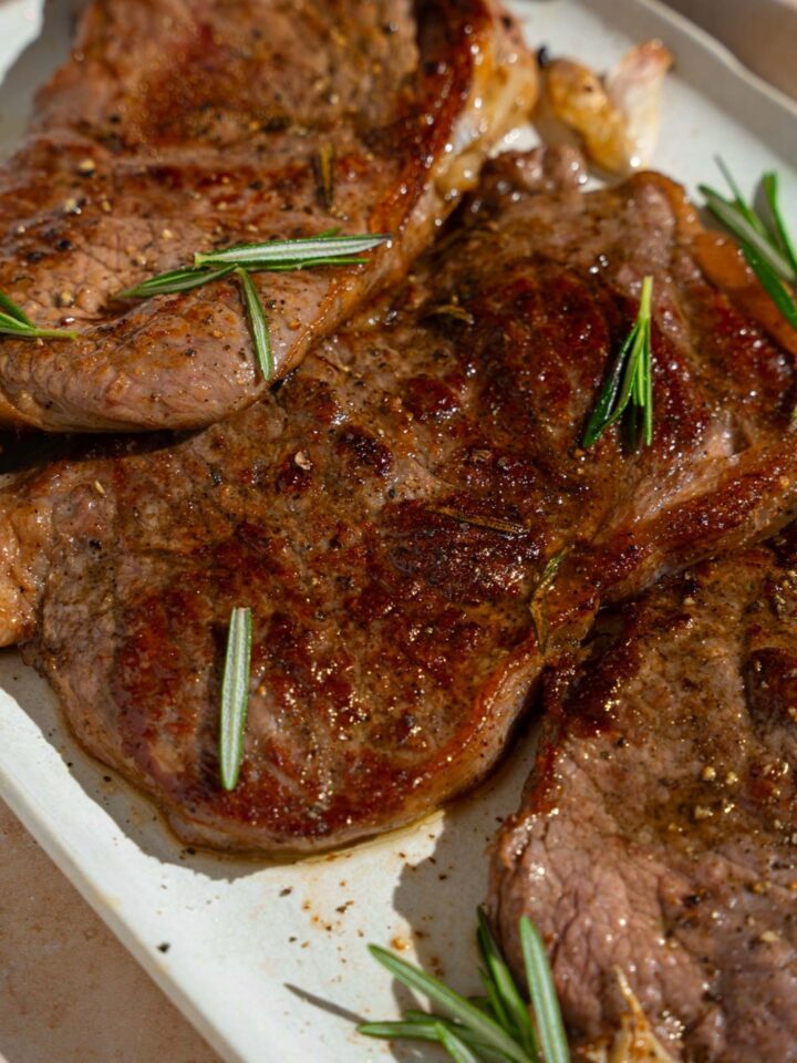 A tray of pan-fried seasoned sirloin steaks. The steaks are on a white platter with fresh rosemary and garlic. The tray is on a tan counter.