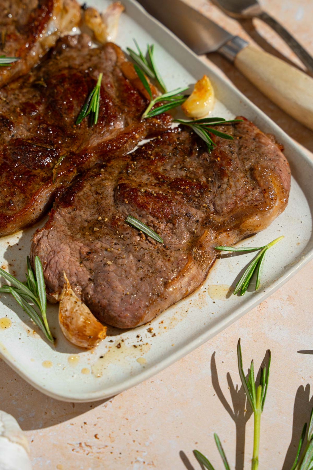 A tray of pan-fried seasoned sirloin steaks. The steaks are on a white platter with fresh rosemary and garlic. The tray is on a tan counter with a knife.