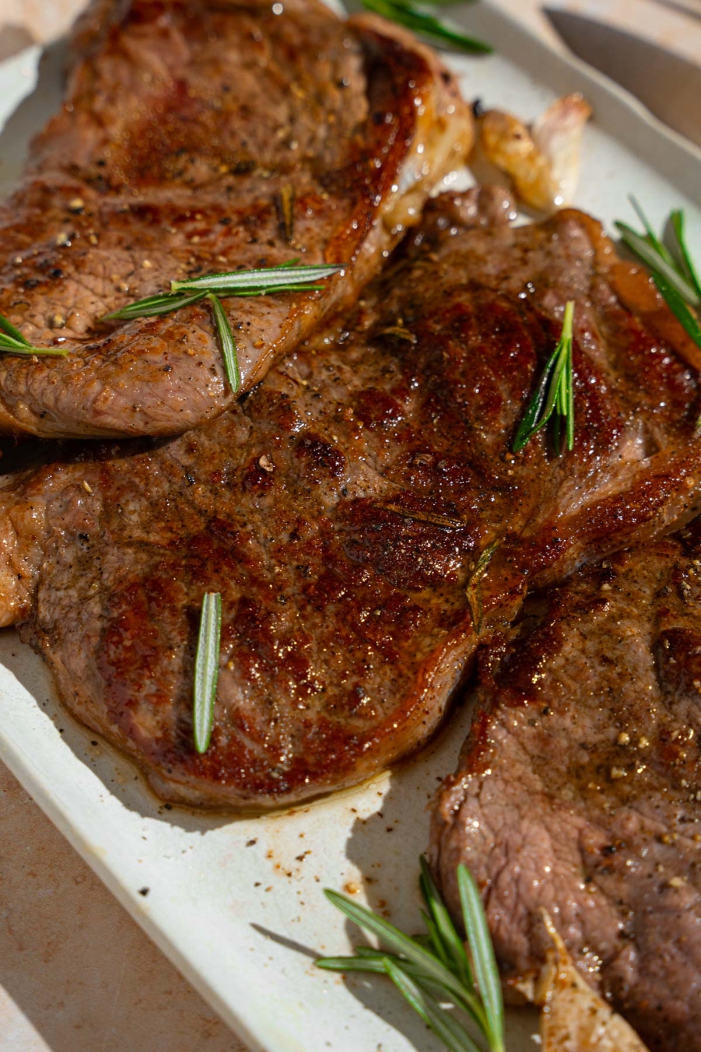 A tray of pan-fried seasoned sirloin steaks. The steaks are on a white platter with fresh rosemary and garlic. The tray is on a tan counter.