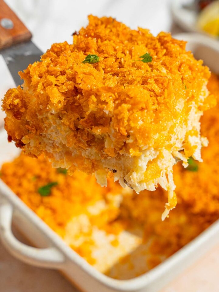 A spatula with a piece of party potatoes casserole with a crispy corn flakes topping garnished with fresh parsley. There is a baking dish with potatoes blurred in the background.