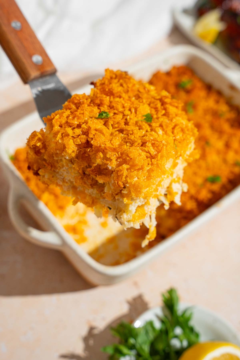 A spatula with a piece of baked potatoes casserole with a crispy corn flakes topping garnished with fresh parsley. There is a baking dish with potatoes and small plate of garnishes blurred in the background.