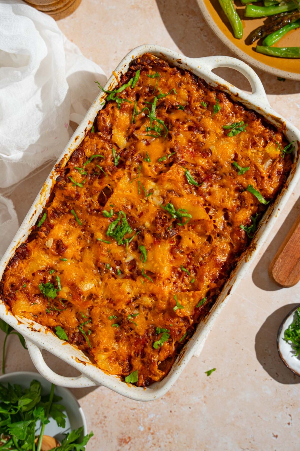 A baking dish with poor man's husband casserole garnished with fresh parsley. The dish is on a tan counter with a plate of garnishes and white cloth napkin.