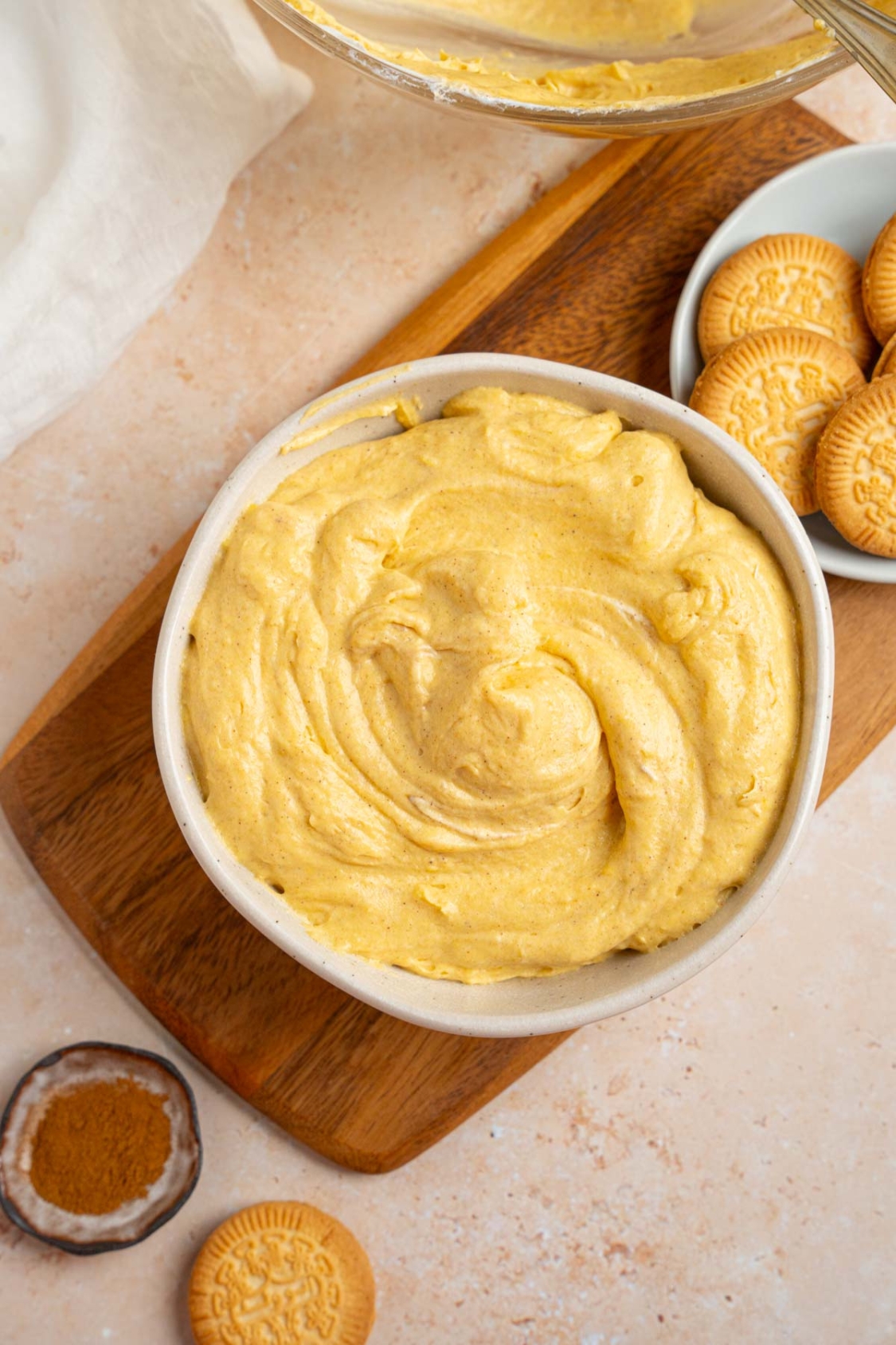 A white bowl with pumpkin fluff. The bowl is on a wooden board with a small bowl of vanilla cookies. The board is on a tan counter with a small ramekin of pumpkin spice and glass bowl of fluff.