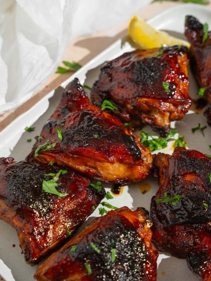 Several sticky chicken thighs served on a white platter garnished with fresh parsley and lemon. The platter is on a tan counter with a white cloth napkin.