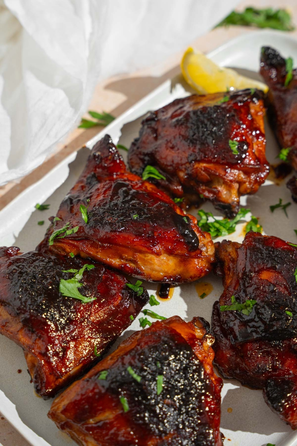 Several sticky chicken thighs served on a white platter garnished with fresh parsley and lemon. The platter is on a tan counter with a white cloth napkin.