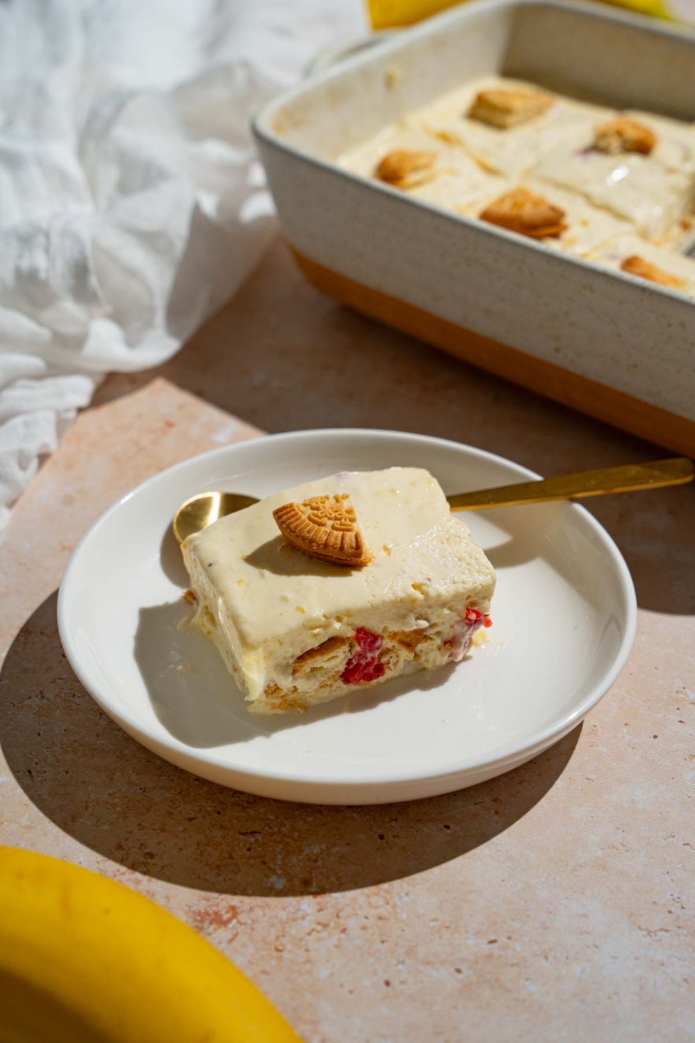 A slice of strawberry banana puding topped with a piece of a vanilla cookie. The pudding is on a white plate with a spoon. The plate is on a tan counter with a baking dish with pudding.