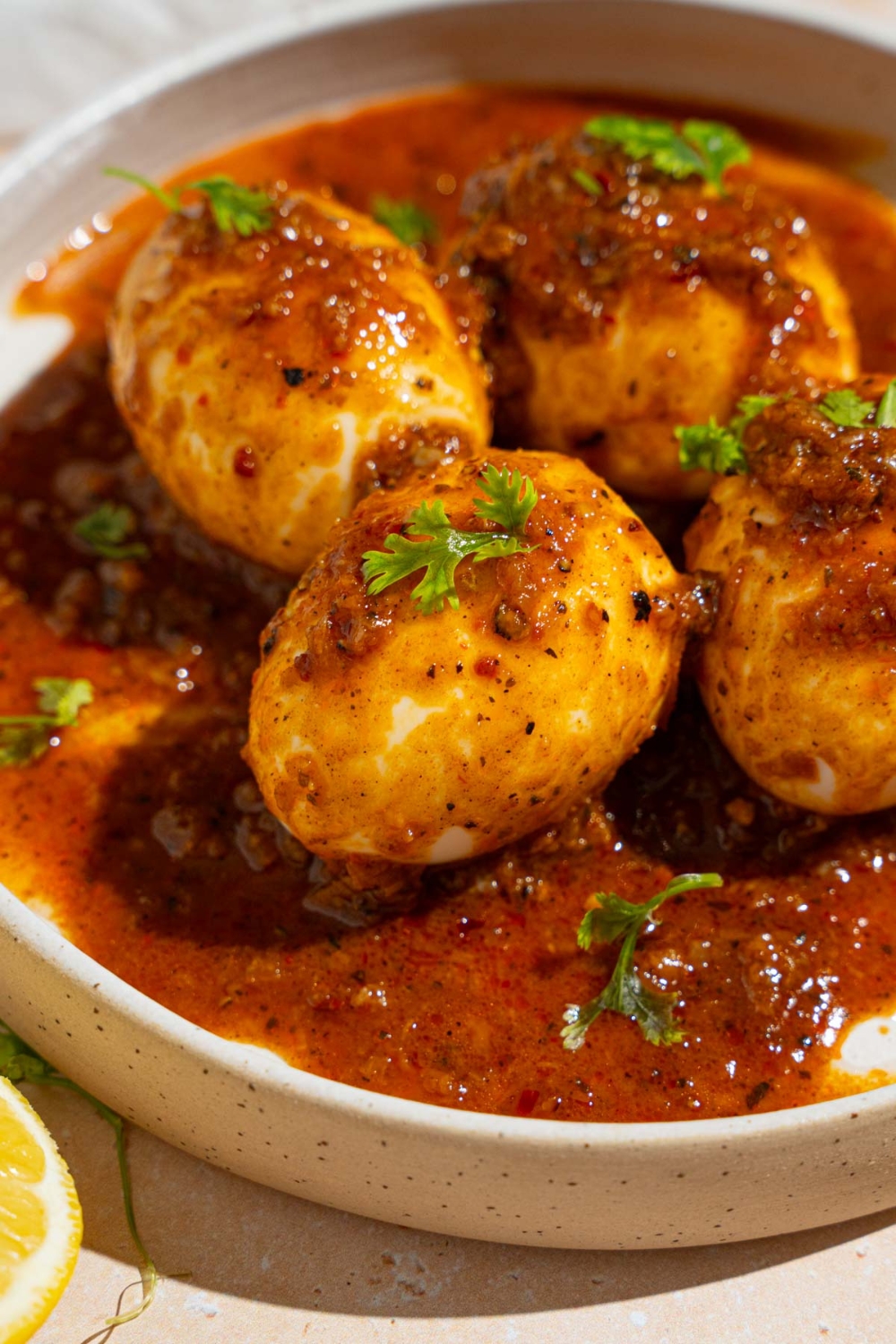 A white plate with four cajun boiled eggs covered in cajun sauce garnished with fresh parsley. The plate is on a tan counter with a sliced lemon.