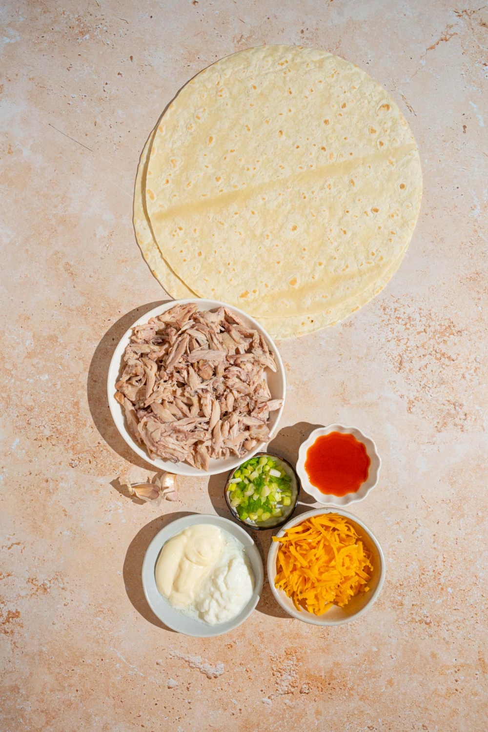 An overhead shot of several bowls in various sizes containing ingredients to make cheesy garlic chicken wraps in including tortilla wraps, shredded chicken, shredded cheddar cheese, sliced green onion, mayo, greek yogurt, and hot sauce.
