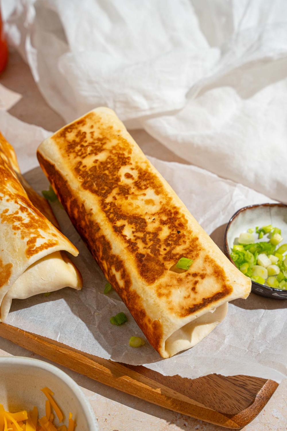 Two cheesy garlic chicken wraps on a wooden board lined with parchment paper. There is a small ramekin of sliced green onion on the board. The board is on a tan counter with a bowl of shredded cheddar cheese and white cloth napkin.