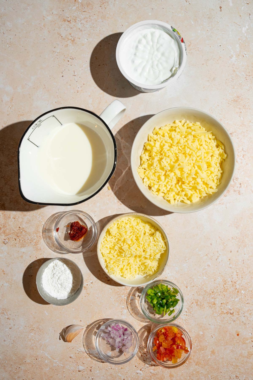 An overhead shot of several bowls in various sizes containing ingredients to make Chipotle queso including milk, sour cream, shredded Monterey jack cheese, onion, chopped peppers, garlic, cornstarch, grated white cheddar, tomatoes, and onion.