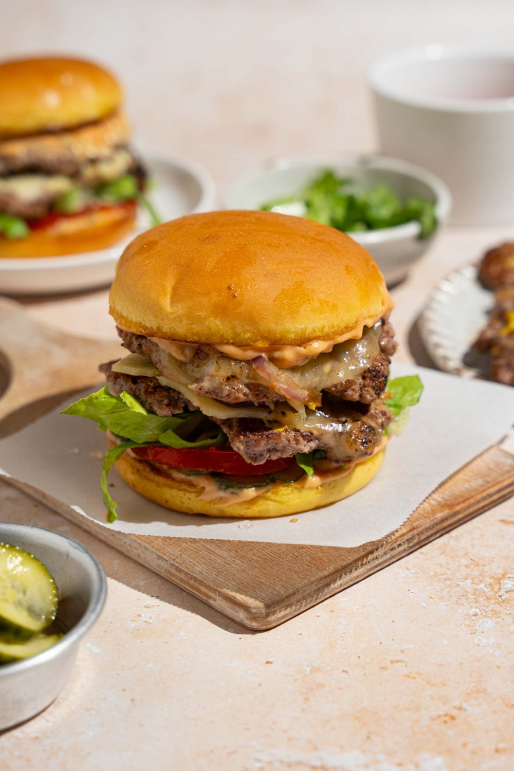 A copycat In-N-Out burger on a wooden board lined with parchment paper. The board is on a tan counter with an additional burger on a plate and small bowl of lettuce.