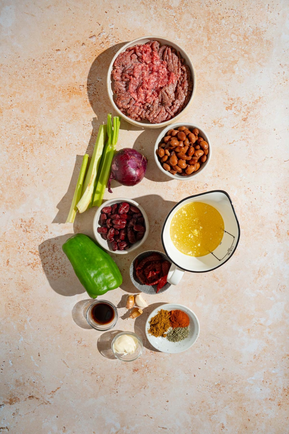 An overhead shot of several bowls in various sizes containing ingredients to make copycat Zippy's chili including ground beef, Portuguese sausage, pinto beans, kidney beans, chicken broth, mayo, red onion, green pepper, celery, and seasonings.