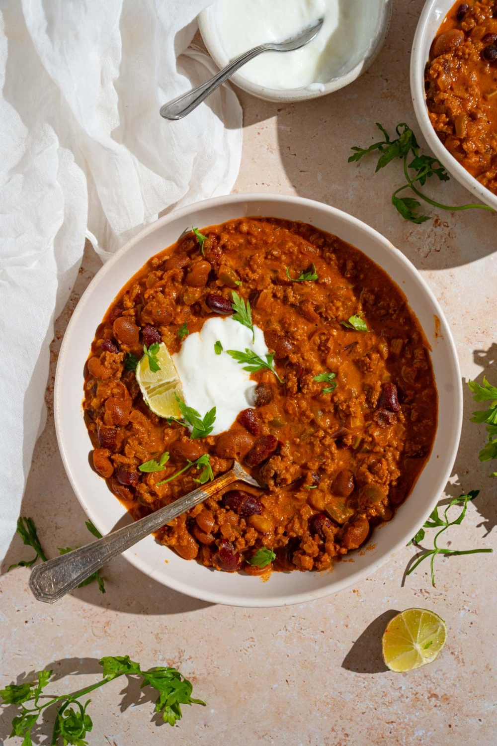 A bowl of copycat Zippy's chili garnished with sour cream, fresh parsley, and lime. There is a spoon in the bowl. The bowl is on a tan counter with a bowl of sour cream, lime, and white cloth napkin.