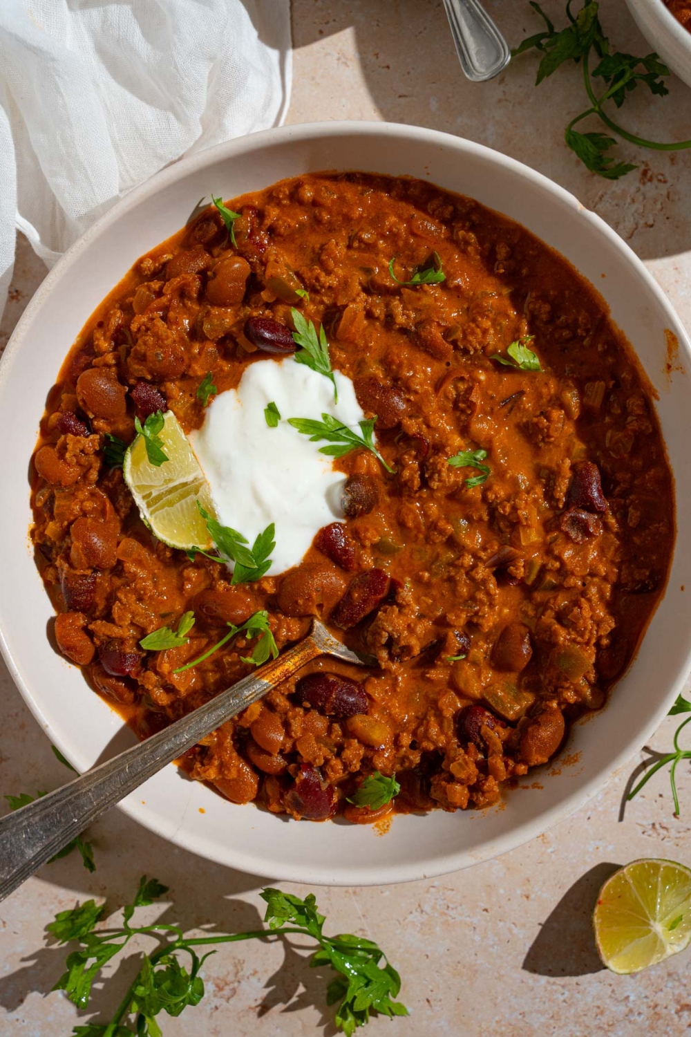 A bowl of copycat Zippy's chili garnished with sour cream, fresh parsley, and lime. There is a spoon in the bowl. The bowl is on a tan counter with lime and white cloth napkin.