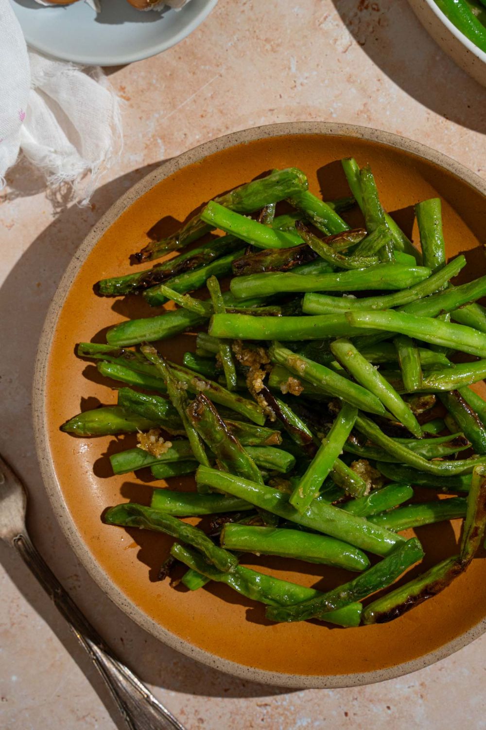 Copycat Din Tai Fung green beans tossed with roasted garlic on an orange plate. The plate is on a tan counter with a fork and white cloth napkin.