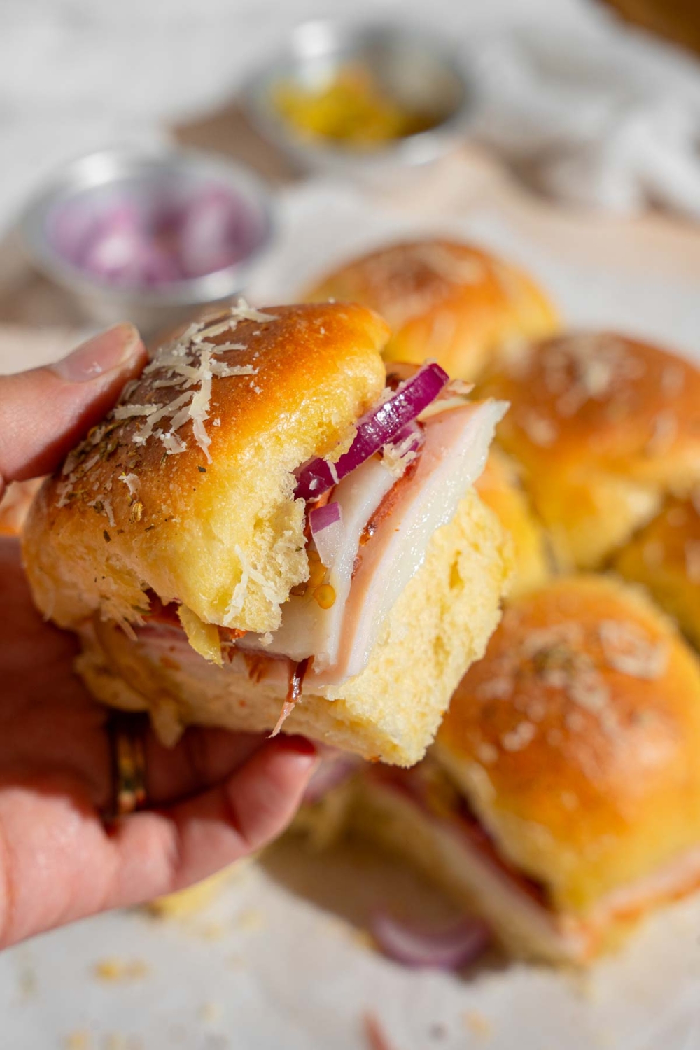 A hand holding an Italian slider served with sliced turkey, cheese, and red onion. There is a tray of sliders lined with parchment paper blurred in the background.