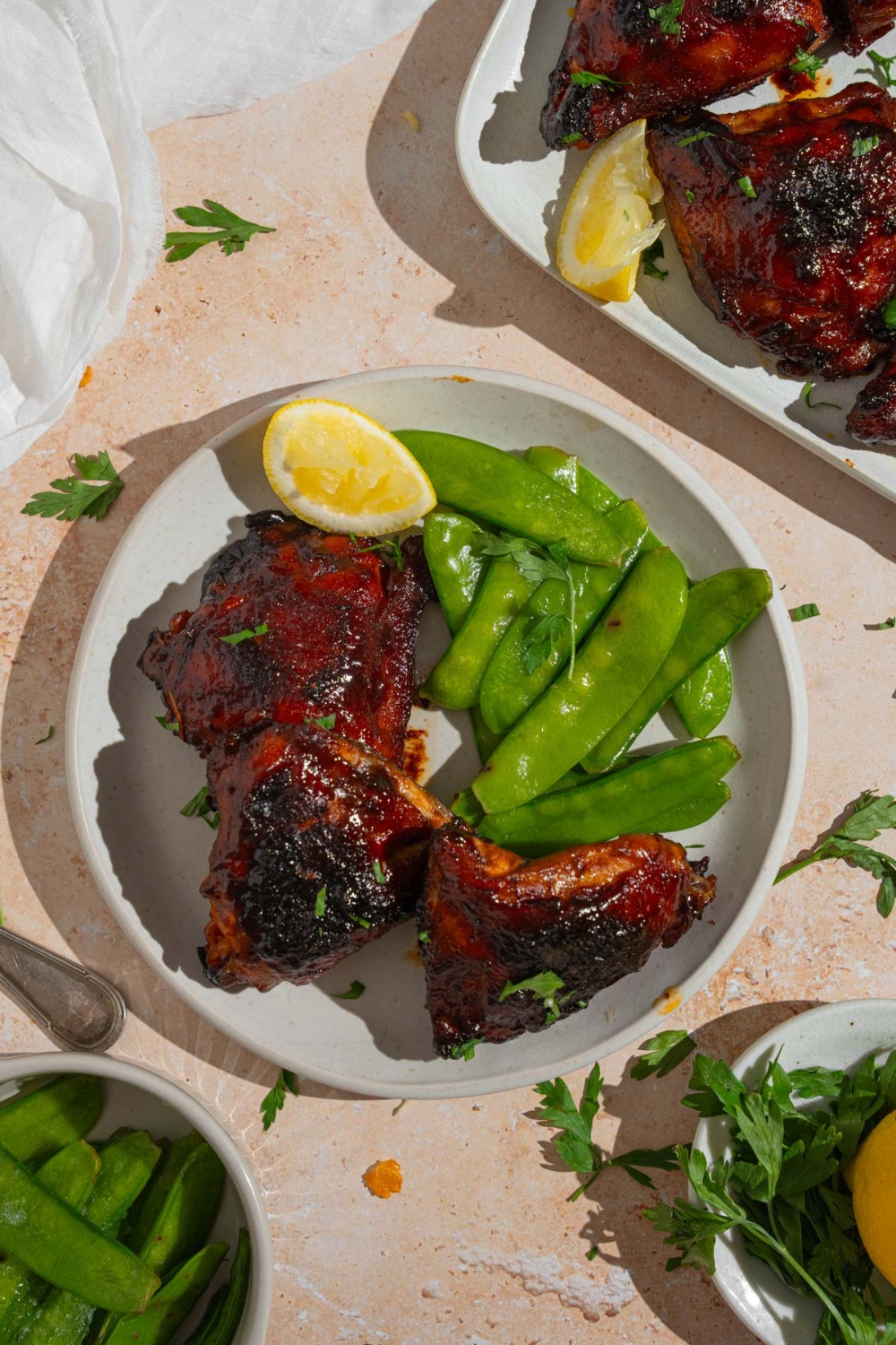 Three sticky chicken thighs served on a white plate with steamed snow peas and a lemon wedge. The plate is on a tan counter with a platter of chicken and small bowls with peas and garnishes.