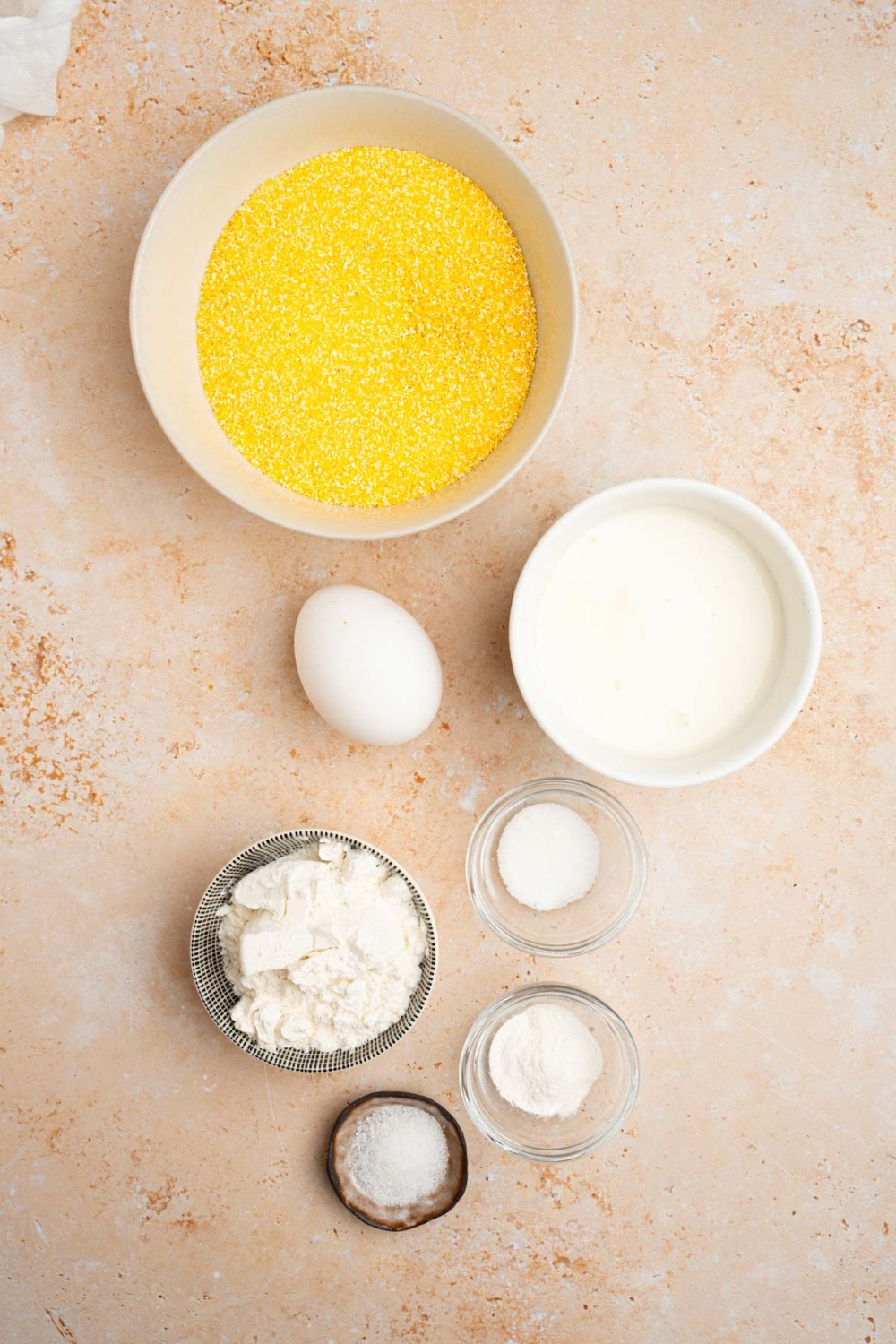 An overhead shot of several bowls in various sizes containing ingredients to make fried cornbread including cornmeal, self-rising flour, milk, sugar, baking powder, egg, and oil.