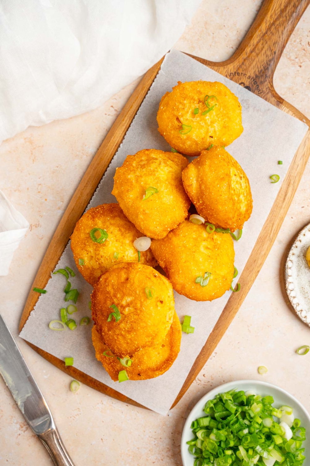 Several pieces of fried cornbread on a wooden board lined with parchment paper and garnished with sliced green onion. The board is on a tan counter with a small bowl of garnishes.