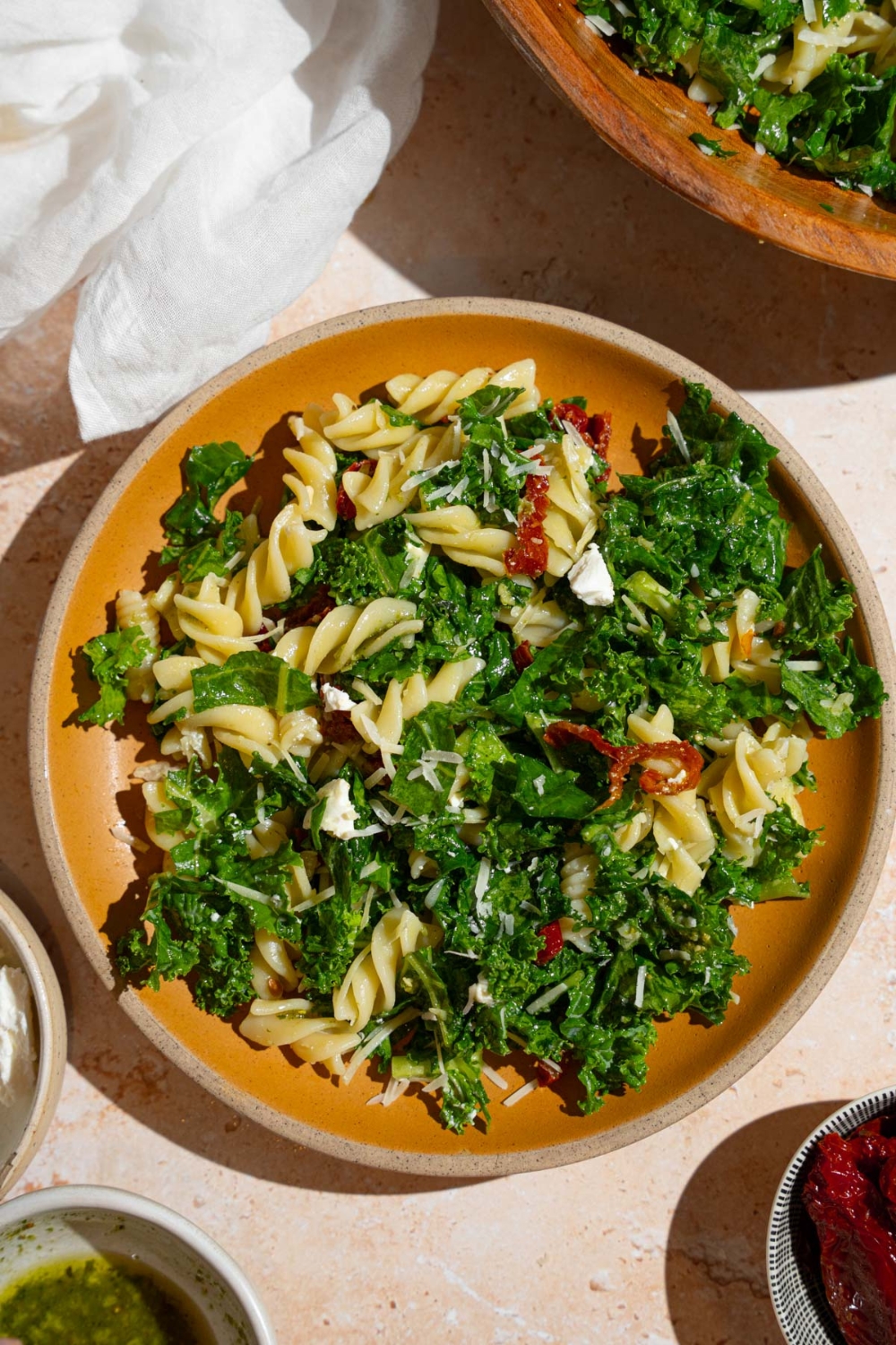 A plate with Mediterranean pasta salad garnished with grated parmesan. The salad consists of kale, rotini pasta, and sundried tomatoes. The plate is on a tan counter with a wooden bowl of pasta salad and white cloth napkin.