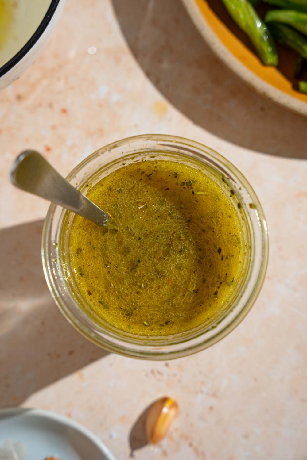A jar of copycat Texas Roadhouse italian dressing with a spoon in the jar. The jar is on a tan counter with a plate of green beans.