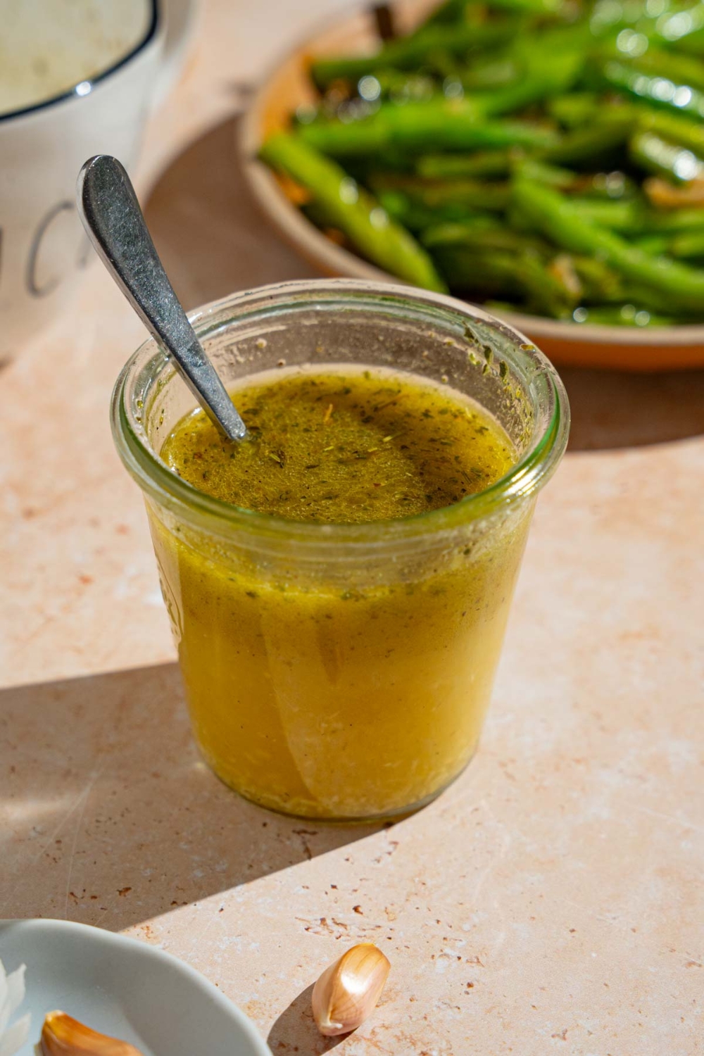 A jar of copycat Texas Roadhouse italian dressing with a spoon in the jar. The jar is on a tan counter with a plate of green beans.