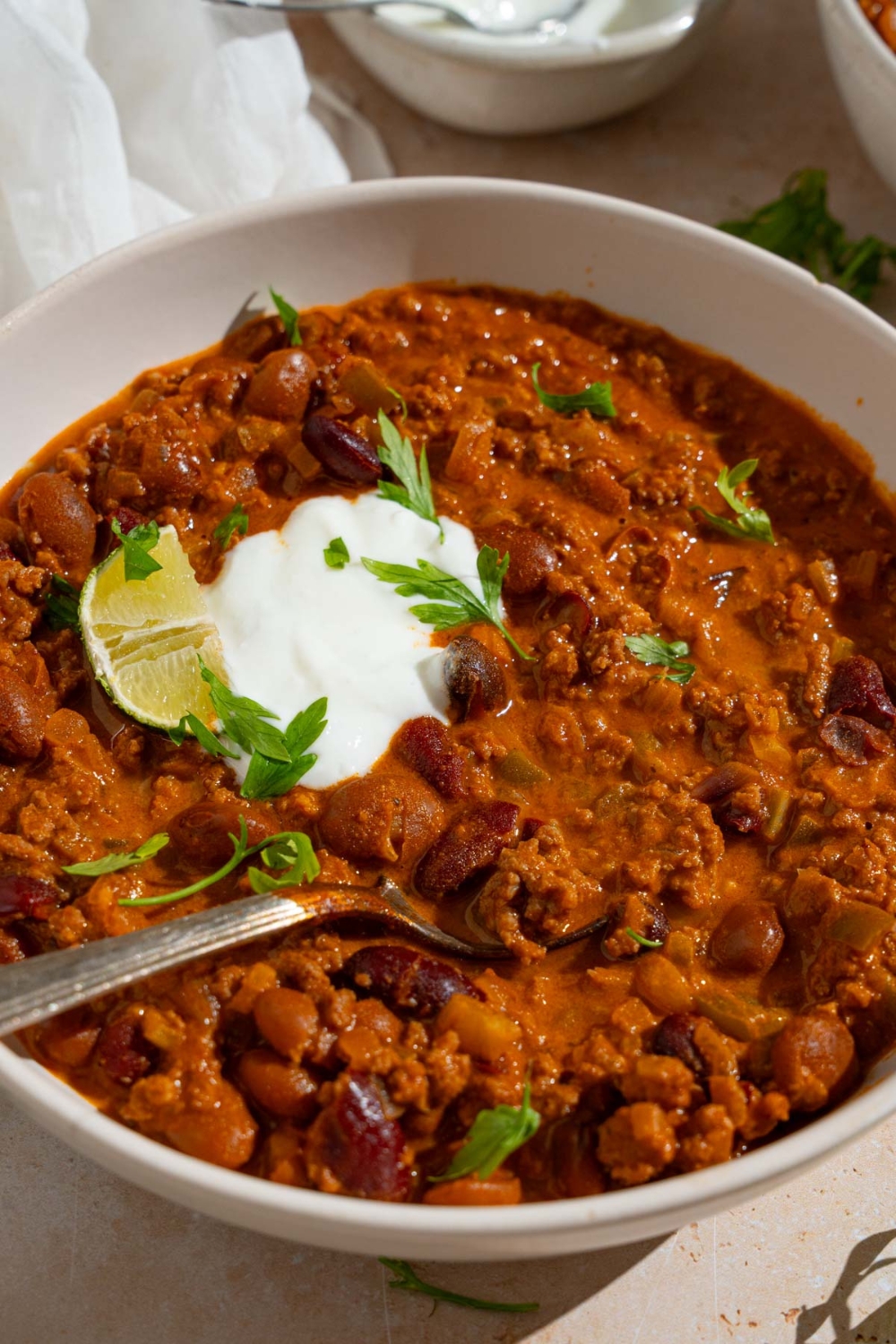 A bowl of copycat Zippy's chili garnished with sour cream, fresh parsley, and lime. There is a spoon in the bowl. The bowl is on a tan counter with a bowl of sour cream and white cloth napkin.