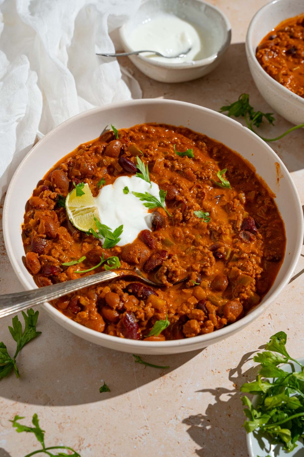 A bowl of copycat Zippy's chili garnished with sour cream, fresh parsley, and lime. There is a spoon in the bowl. The bowl is on a tan counter with a bowl of sour cream and white cloth napkin.