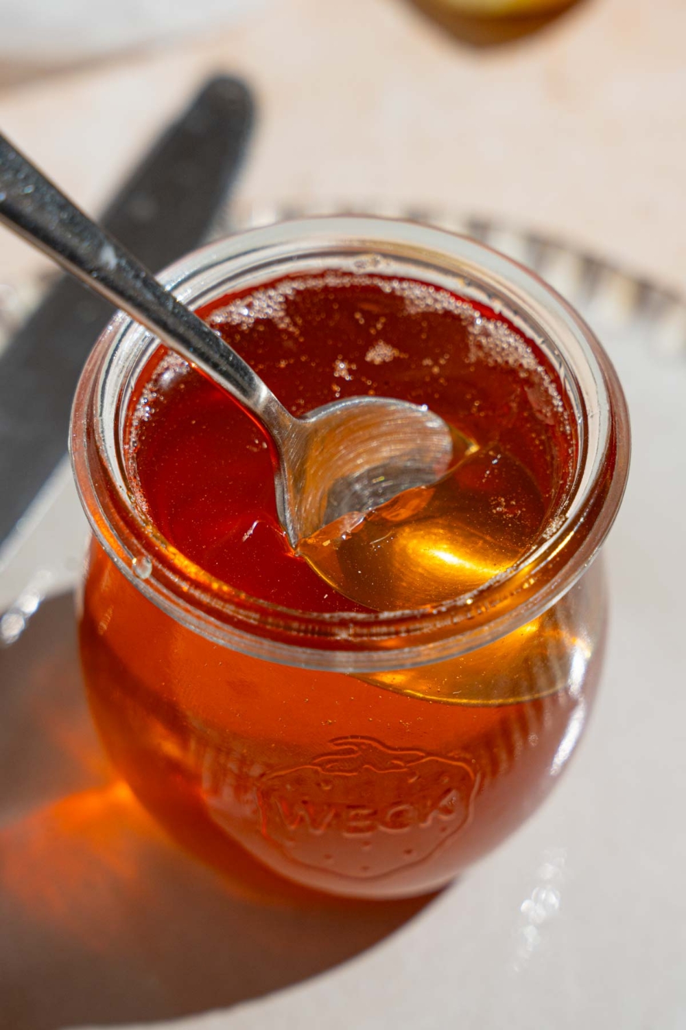 A glass jar of apple jelly with a spoon in the jelly. The jar is on a plate on a tan counter with a knife.