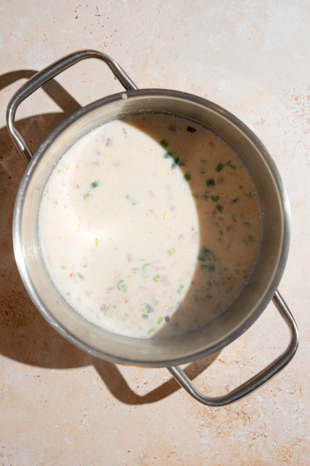 A stock pot with heated milk with chopped peppers, onion, and garlic. The pot is on a tan counter.