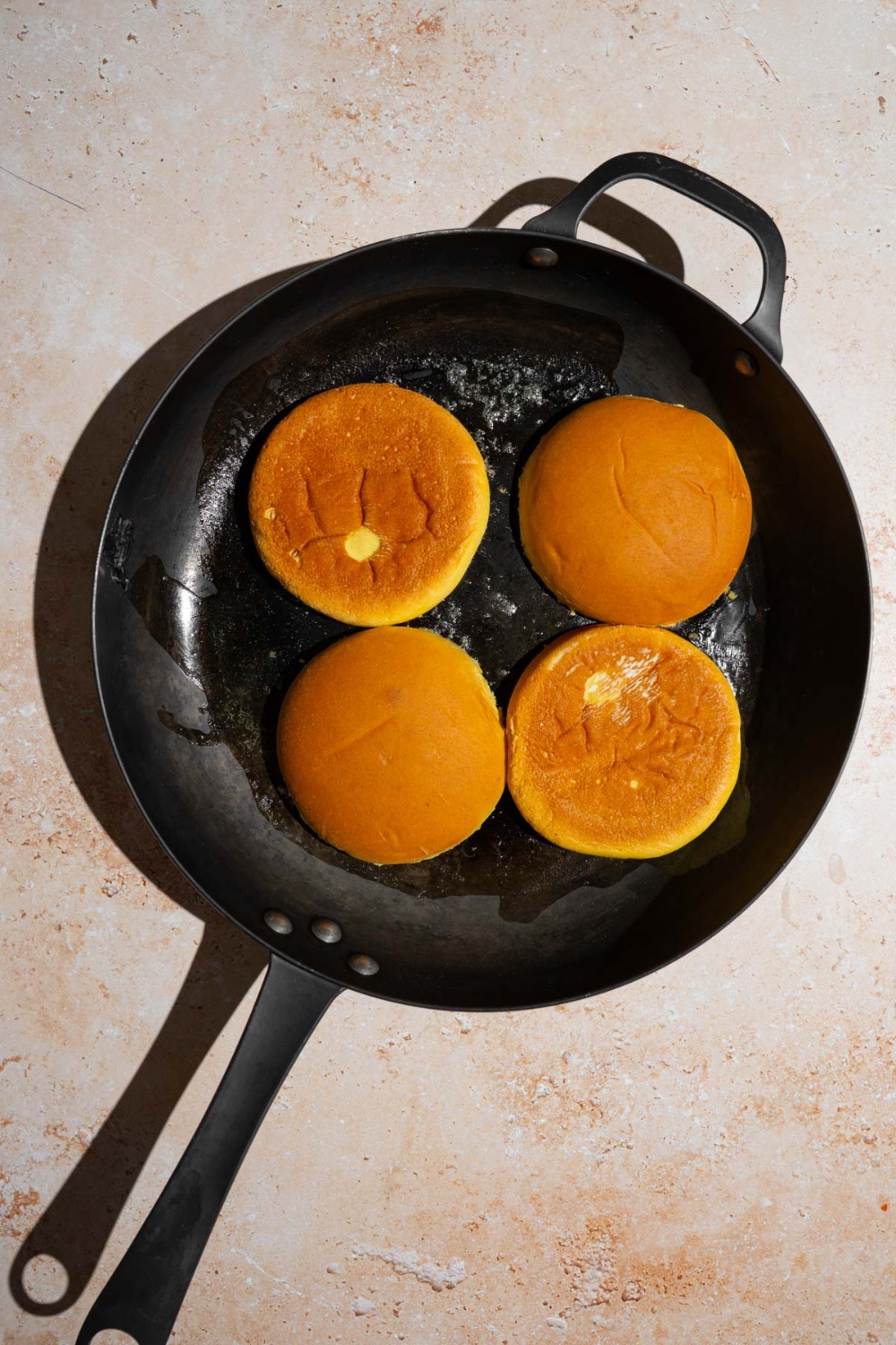 A skillet with two burger buns toasting with butter. The skillet is on a tan counter.