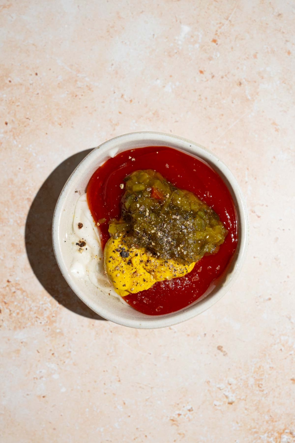 A white bowl with ingredients to make In-n-Out sauce including ketchup, mayo, mustard, relish, and seasonings. The bowl is on a tan counter.