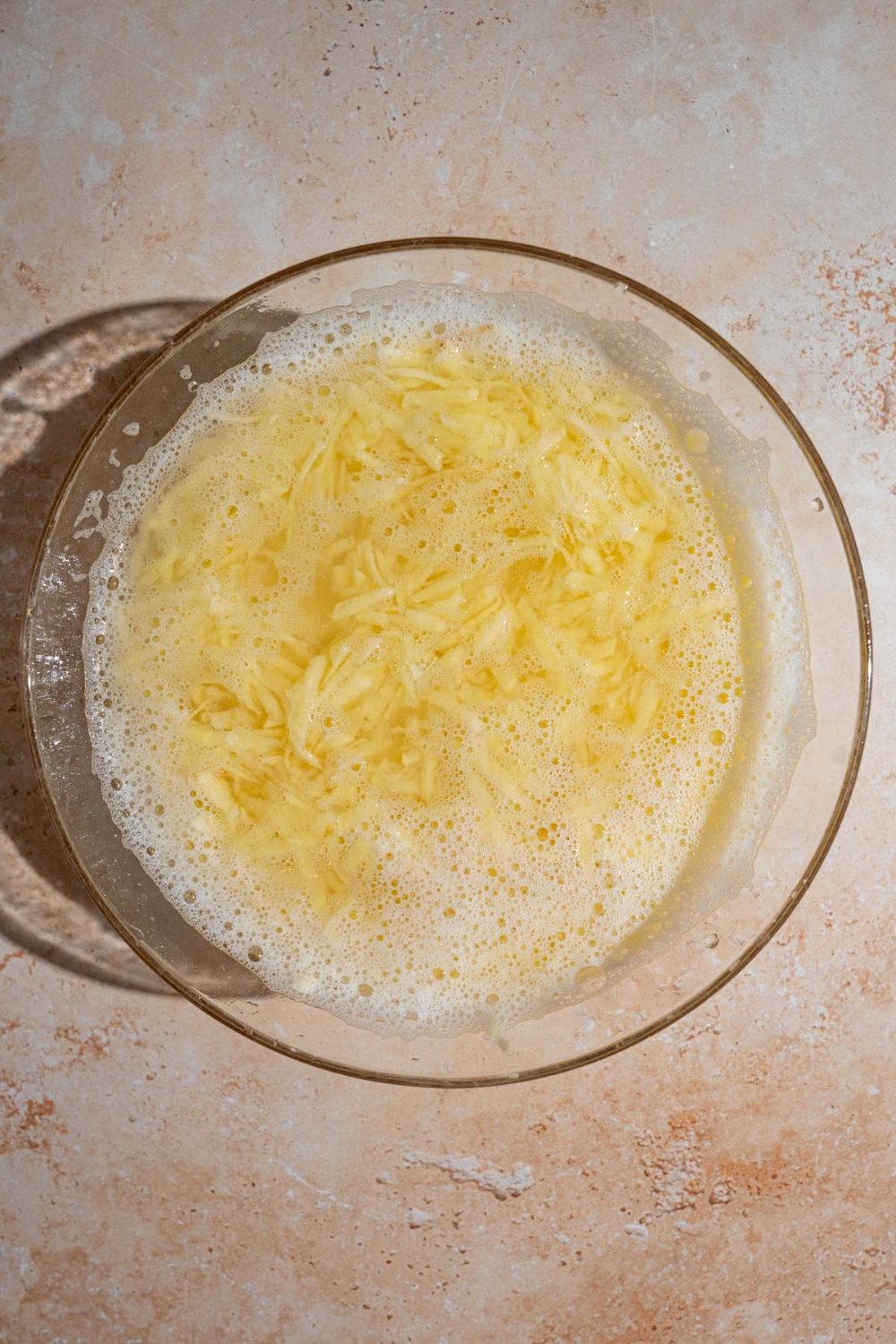 A bowl of shredded potatoes soaking in water. The bowl is on a tan counter.