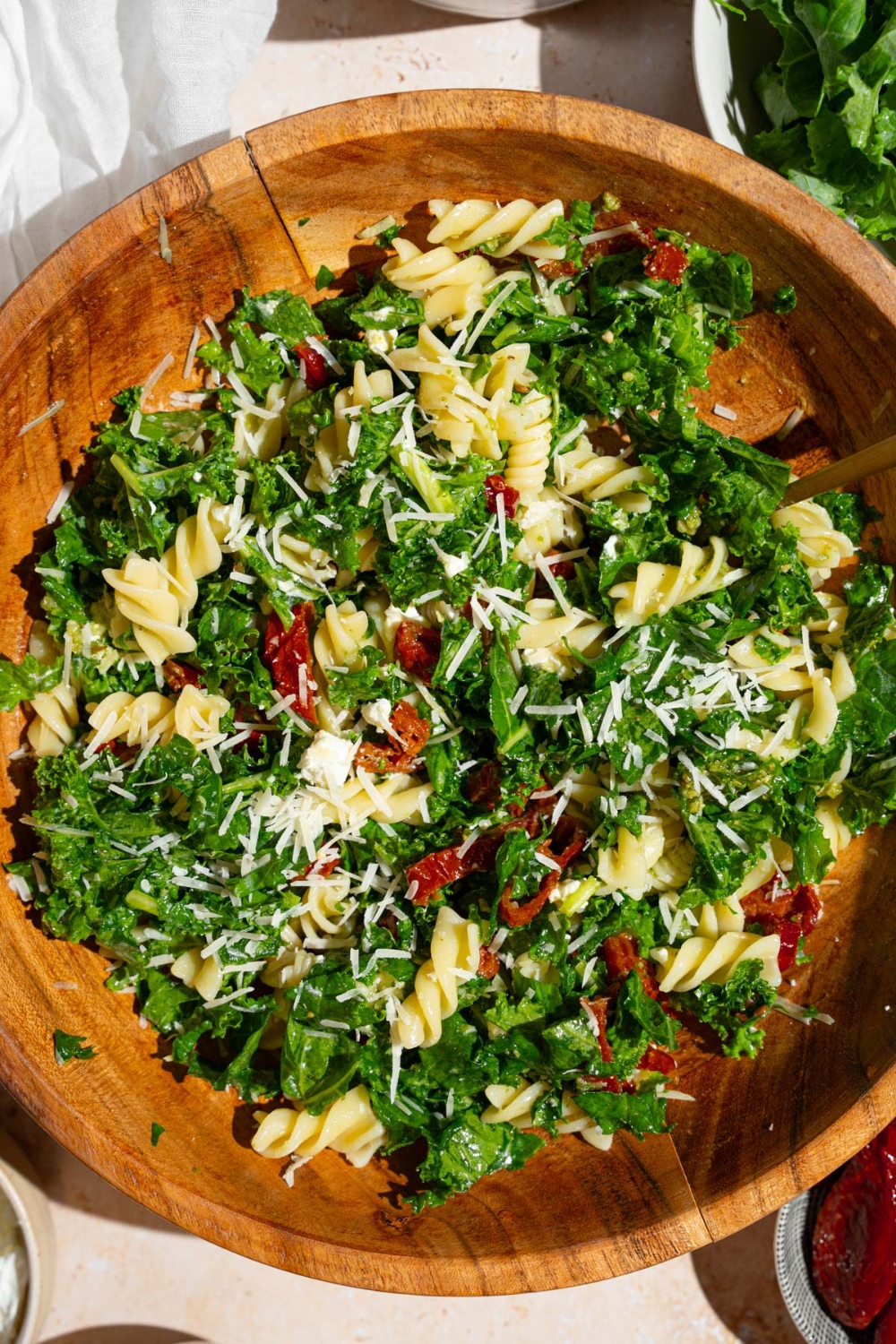 Mediterranean pasta salad served in a wooden bowl garnished with grated parmesan. The salad includes kale, rotini pasta, and sundried tomatoes. There is a fork in the bowl.
