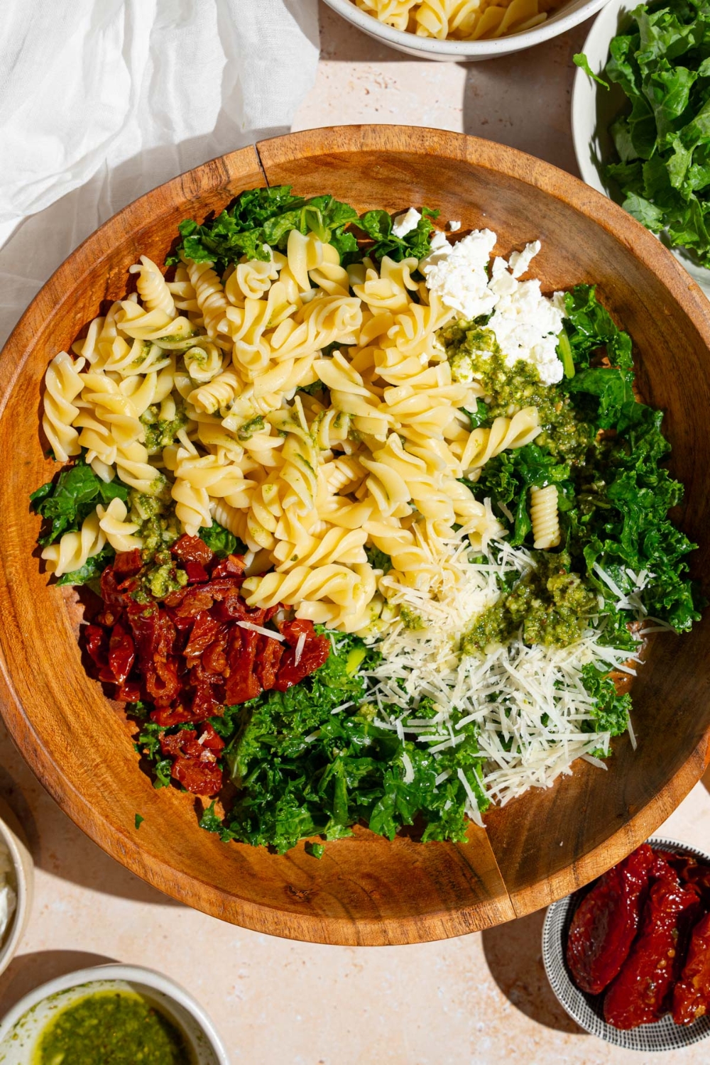 A wooden bowl with ingredients to make Mediterranean pasta salad including kale, rotini pasta, sundried tomatoes, feta cheese, and grated parmesan. The bowl is on a tan counter with a white cloth napkin and bowls with salad ingredients.