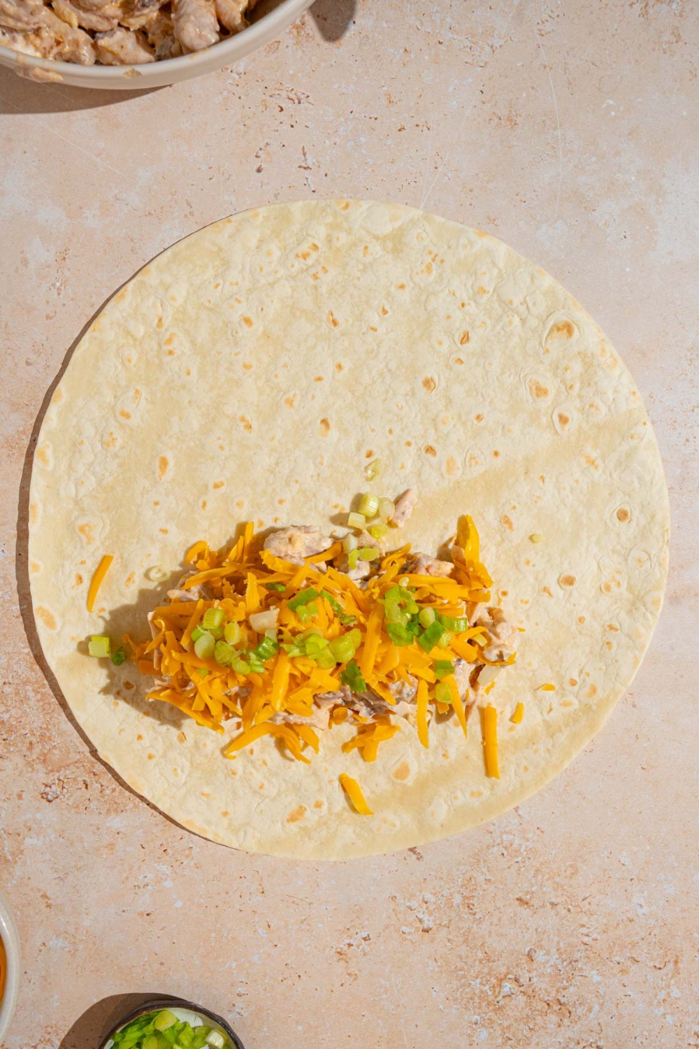 An open tortilla wrap with a garlic chicken mixture topped with shredded cheese and sliced green onion. The wrap is on a tan counter.