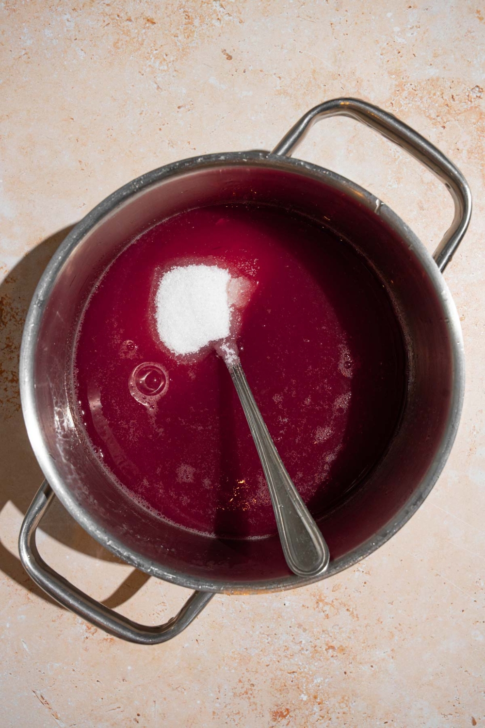 A stock pot with cooked chokecherry jelly with a spoon stirring in sugar. The pot is on a tan counter.