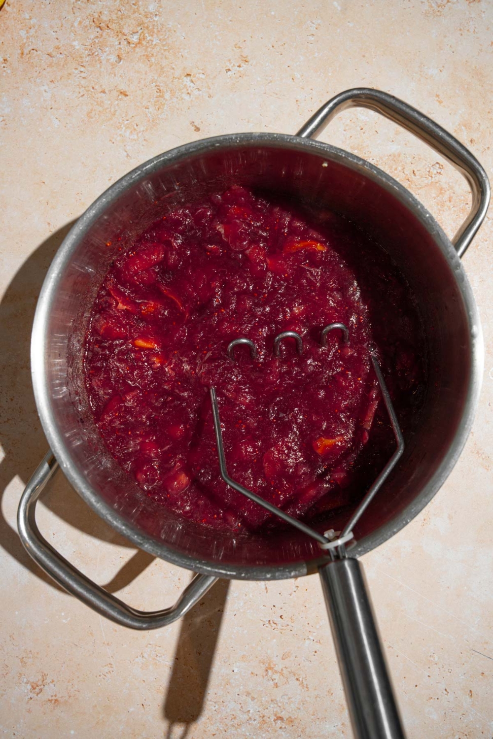 A stock pot with a potato masher mashing chokecherry jelly mixture. The pot is on a tan counter.