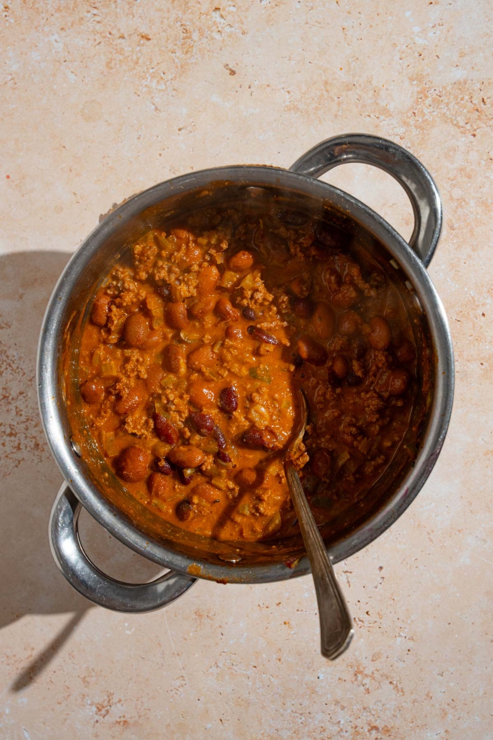 A stock pot with a spoon stirring cooked copycat Zippy's chili. The pot is on a tan counter.