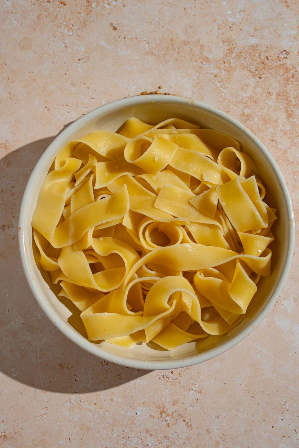 A bowl of cooked wide egg noddles. The bowl is on a tan counter.