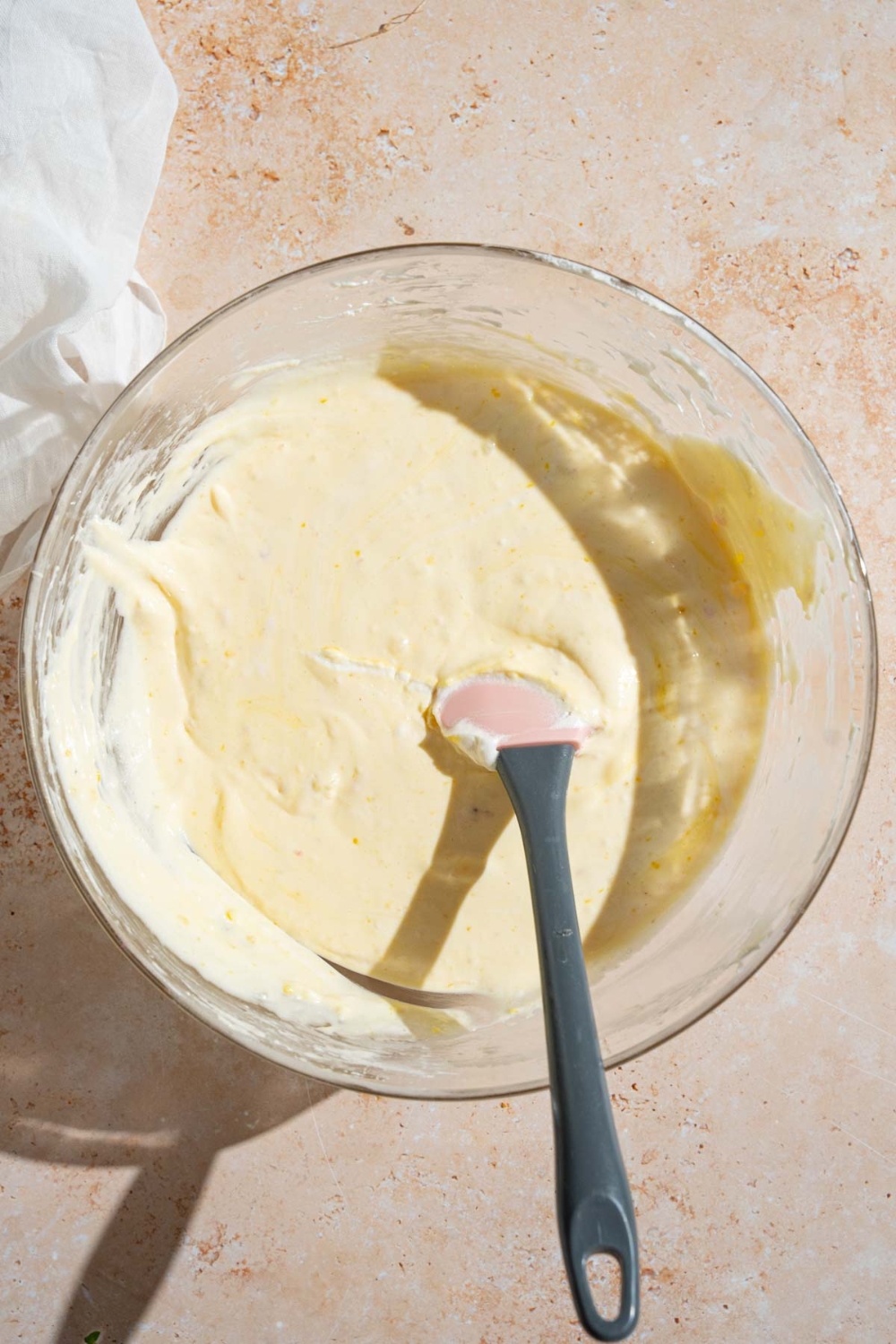 A glass bowl with a spatula mixing a banana pudding mixture. The bowl is on a tan counter with a white cloth napkin.