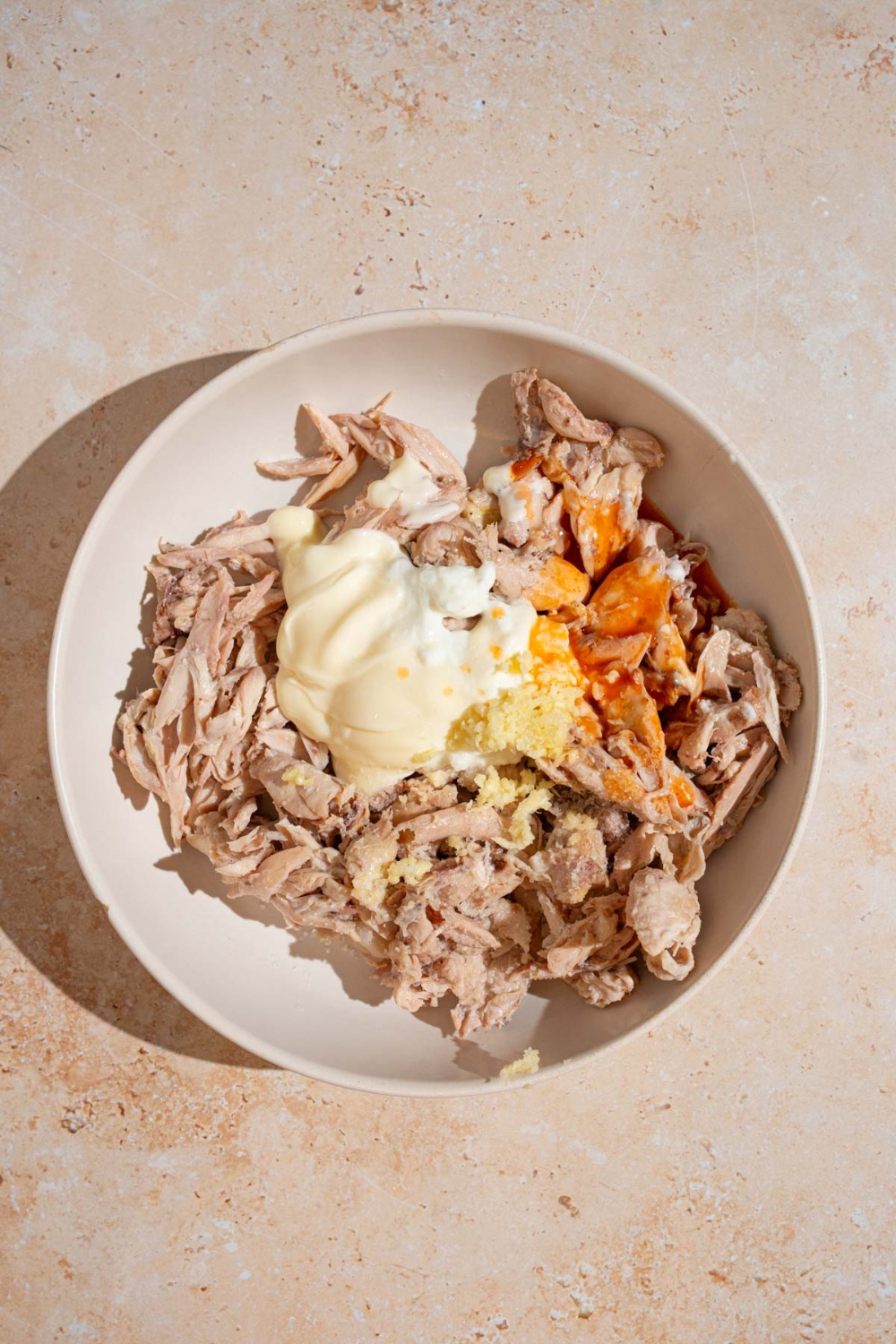 A bowl of shredded chicken, mayo, greek yogurt, and hot sauce. The bowl is on a tan counter.