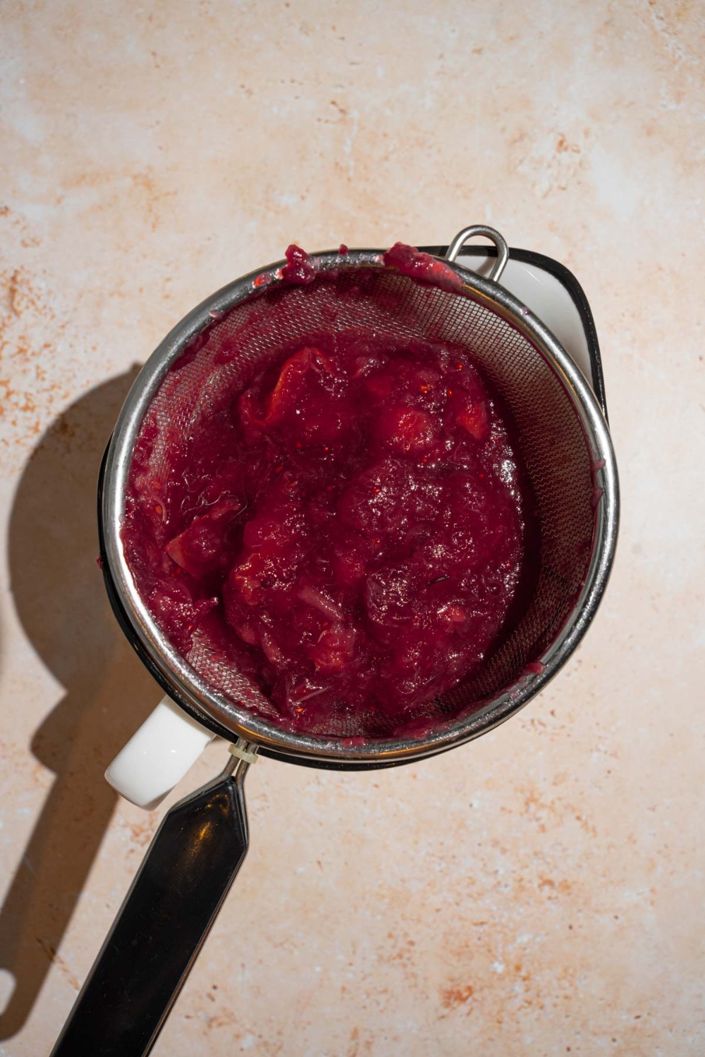 A mesh strainer with strained chokecherry mixture. The strainer is on a tan counter.