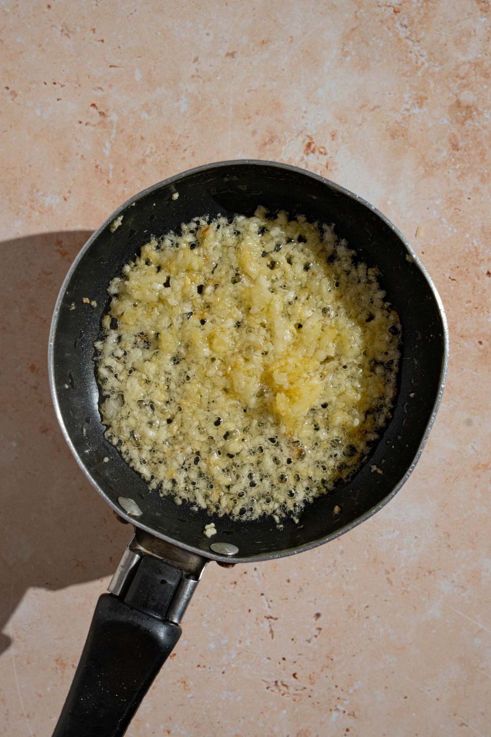 A skillet with chopped garlic cooking in oil. The skillet is on a tan counter.