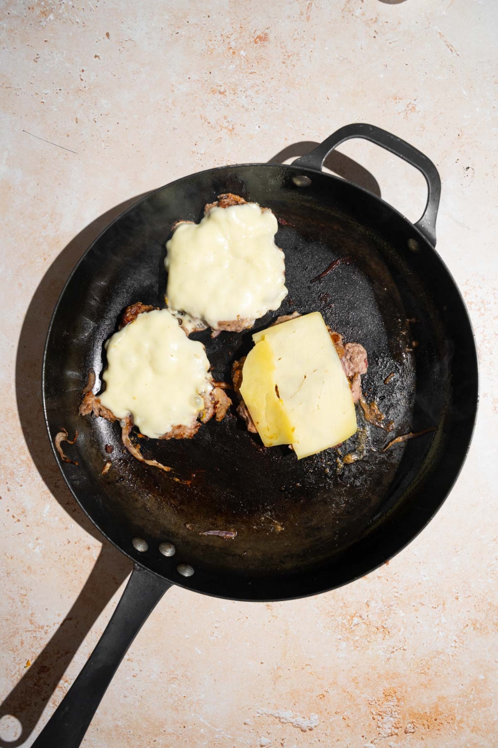 A skillet with three burger patties cooking with mustard and American cheese. The skillet is on a tan counter.