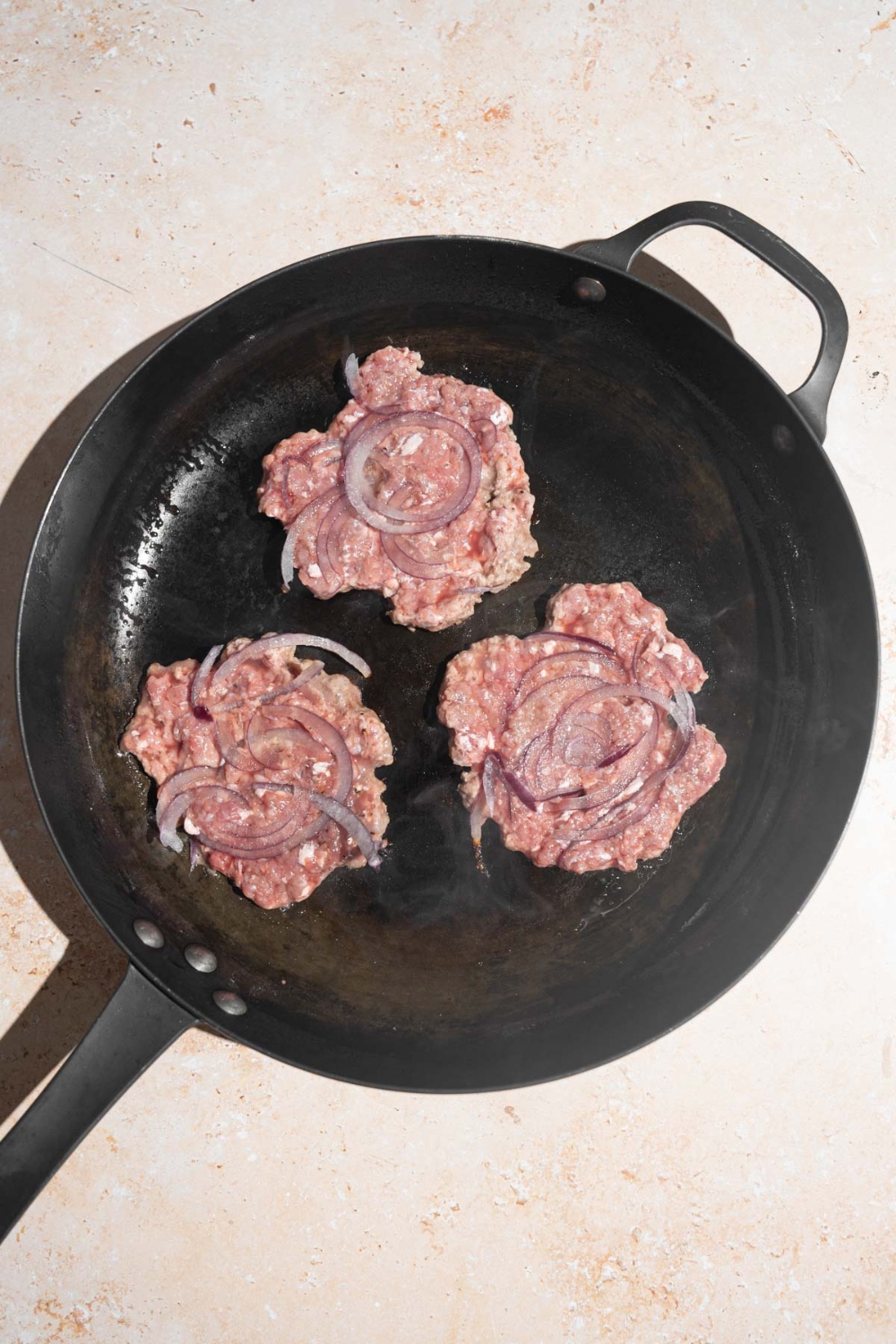 A skillet with three patties cooking with red onion. The skillet is on a tan counter.
