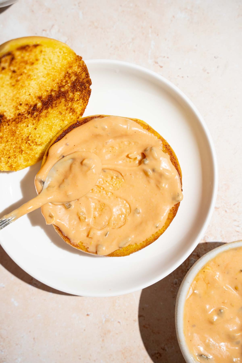 An open toasted bun with a spoon spreading copycat In-n-Out sauce on the bun. The bun is on a white plate. The plate is on a tan counter.