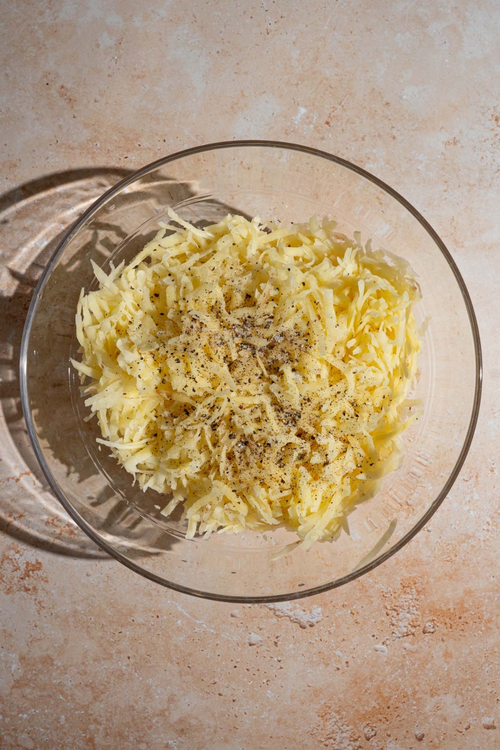 A bowl of shredded potatoes seasoned with salt and pepper. The bowl is on a tan counter.